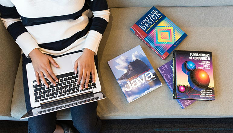 Person using a laptop surrounded by computer science textbooks, including Compiler Design, Java, C++, and Fundamentals of Computing II.
